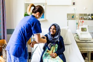 A female patient wearing a headscarf is sitting in a hospital bed smiles up at a female Mercy Health worker in all blue while she takes her blood pressure.