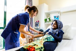A female patient wearing a headscarf lies back in a hospital bed smiling up at a female Mercy Health worker in all blue while she takes her blood pressure.