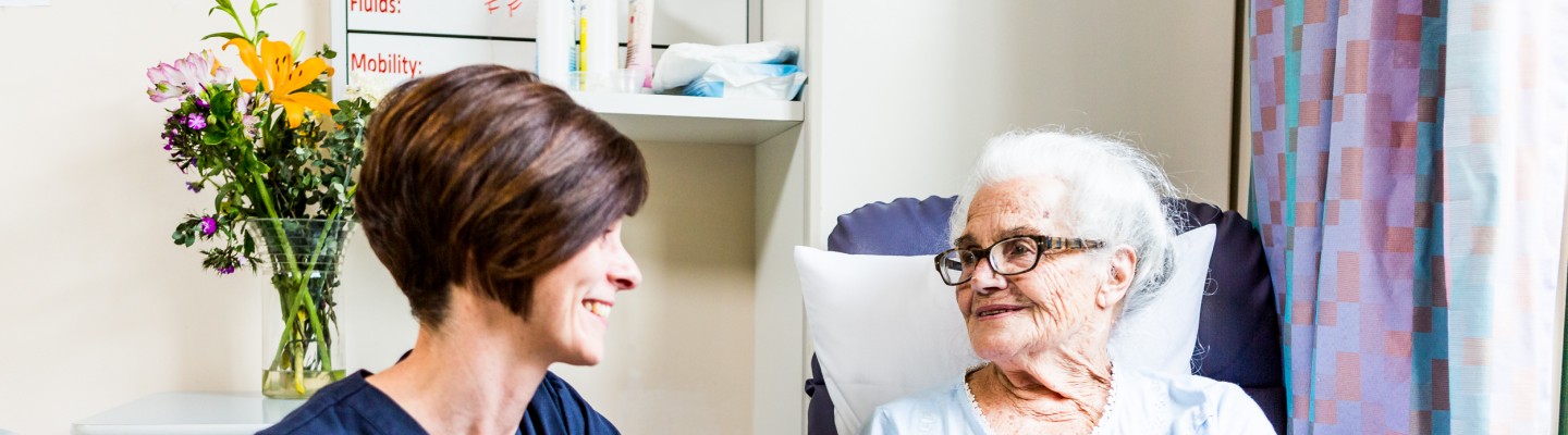 A Mercy Health female worker with short brown hair sits to the left of an elderly resident who holds her hand. They smile at each other.