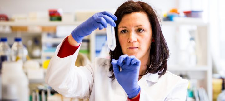 A medium close up of a woman in a lab wearing blue gloves and white lab coat holding a test tube.