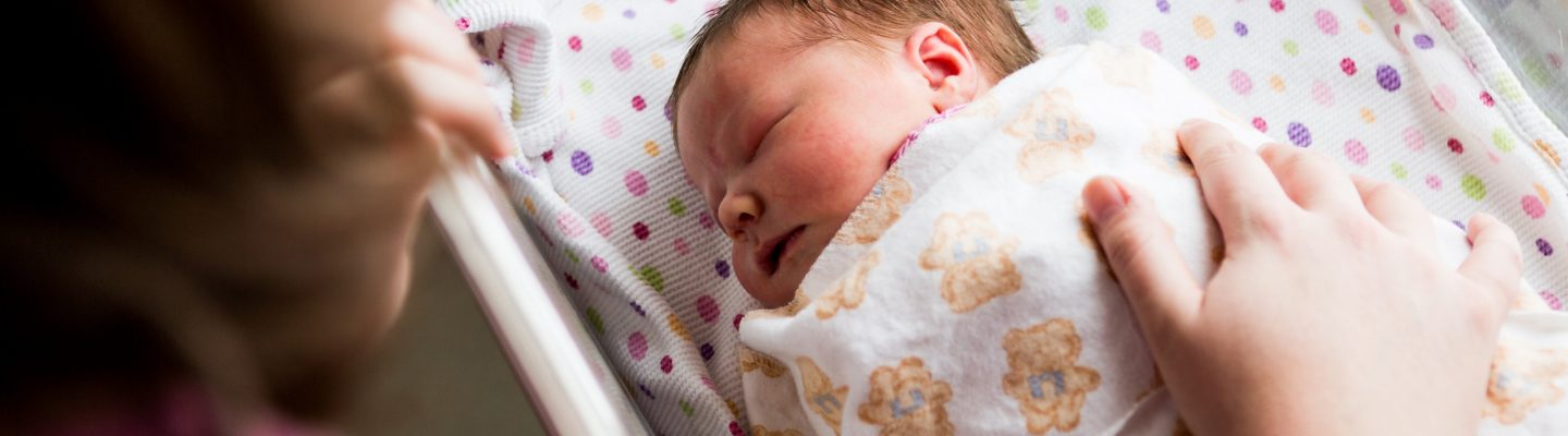 A close up looking down at a new born baby wrapped up and asleep while a woman rests her hand on the baby.