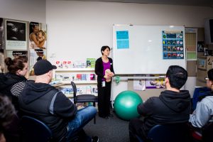 A long shot of a woman standing next to a green yoga ball in front of a room of people sitting and watching her.