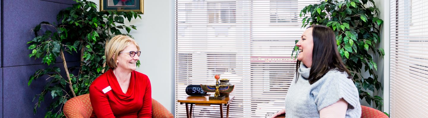 Two women sit in red arm chairs in a small room talking and laughing.