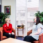 Two women sit in red arm chairs in a small room talking and laughing.