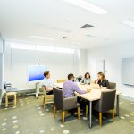 Two men and two women in casual corporate clothing sit at a board table in a white room having a meeting.