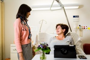 A female patient sits in bed in front of flowers and a propped up tablet, looking up at a female doctor in casual clothing, smiling.